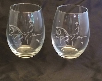 May include: Two clear glass wine glasses with an etched design of a person riding a horse. The glasses are empty and sitting on a dark surface.