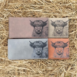 May include: Four leather wallets in shades of pink, beige, gray, and orange, each featuring a detailed illustration of a Highland cow. The wallets are rectangular and appear to be closed, with the cow's face prominently displayed.