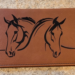 May include: A brown leather checkbook cover with a black silhouette of two horses' heads facing each other.