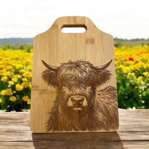 May include: A brown wooden cutting board with a handle features a laser-engraved image of a Highland cow with long, shaggy hair. The cow's face is looking directly at the viewer.