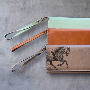 Puede incluir: Tres carteras de cuero de diferentes colores con correas de muñeca. Las carteras están apiladas una encima de la otra. La cartera superior es beige con una imagen en blanco y negro de un caballo. La cartera del medio es marrón. La cartera inferior es verde claro.