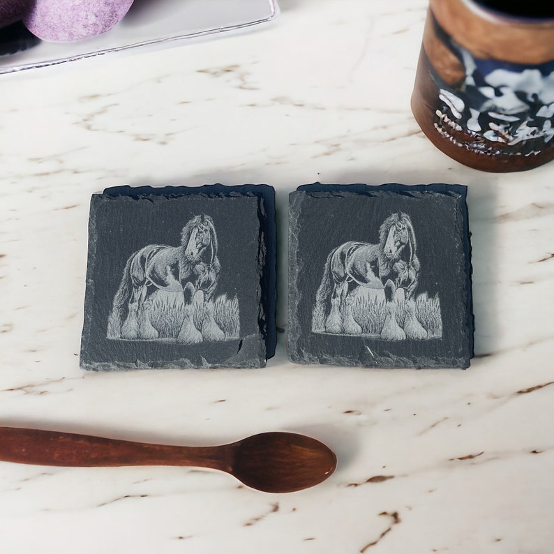 May include: Two black slate coasters with an etched image of a black horse with a white mane and tail. The horse is standing in a field of grass.