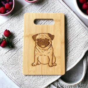 May include: A brown bamboo cutting board with a carved image of a pug dog. The board has a handle cutout.