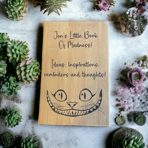 May include: A wooden journal with the text "Jen's Little Book of Madness! Ideas, inspirations, reminders and thoughts!" and a black and white illustration of the Cheshire Cat from Alice in Wonderland.