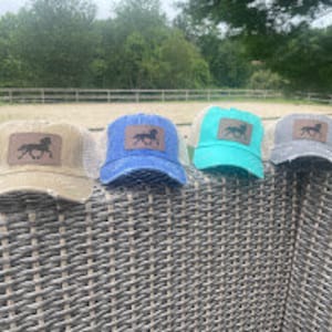 May include: Four trucker hats with a distressed look. The hats are in beige, blue, turquoise, and gray. Each hat has a brown horse silhouette patch on the front.