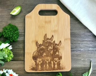 May include: A brown bamboo cutting board with a handle features a laser-engraved image of three donkeys standing close together. The donkeys are brown and white with black eyes and noses.