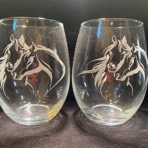 May include: Two clear glass wine glasses with an etched design of two horses' heads facing each other.