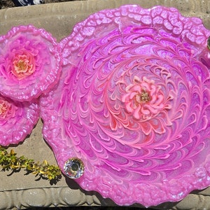 May include: A set of three pink resin coasters with a swirl design and a flower in the center. The coasters are decorated with iridescent glitter and have a scalloped edge.