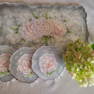May include: A set of four gray resin coasters with a floral design featuring pink peonies. The coasters are set on a gray resin tray with a similar floral design.