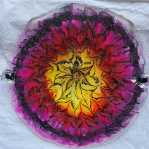 May include: A round resin tray with a colorful abstract design. The design features a yellow, orange, and red center with a black swirl. The center is surrounded by a ring of pink and purple. The tray has a clear, glossy finish.