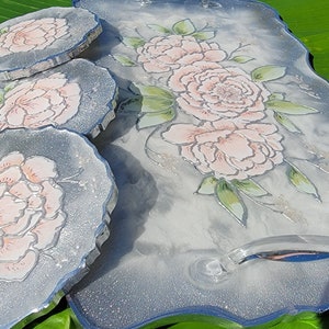 May include: A set of three silver-rimmed coasters with a white background and pink floral designs. The coasters are paired with a larger matching tray with a silver rim and a handle.