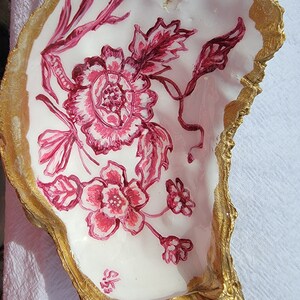 May include: A white oyster shell with a gold rim, painted with a pink floral design. The design features a large pink flower with smaller pink flowers and leaves.