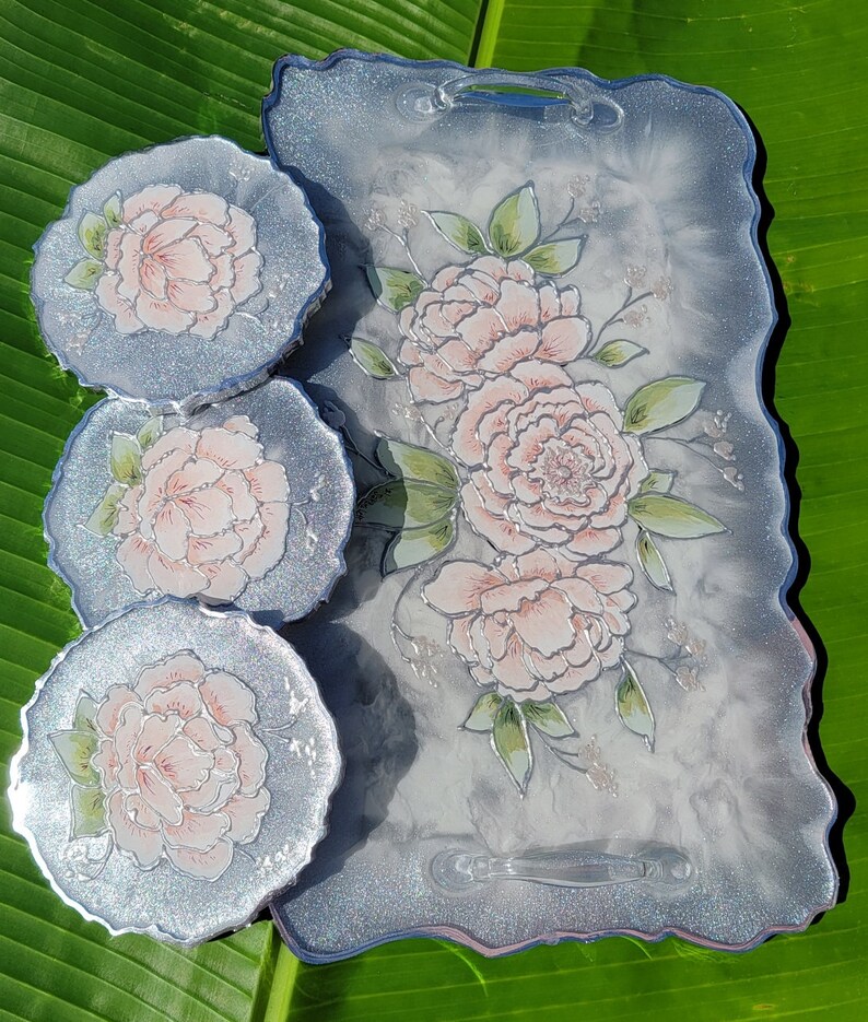 May include: A set of four silver and white coasters with a floral design. The coasters feature a delicate pink flower with green leaves. The coasters are set on a larger silver and white tray with a similar floral design.