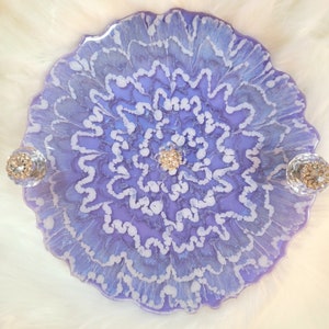 May include: A round, purple and white resin tray with a swirling, floral design. The tray has two decorative knobs on the sides.