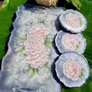 May include: A set of three silver glitter resin coasters with a white background and hand-painted pink peonies. The coasters are round and have a scalloped edge. The tray is also silver glitter resin with a white background and hand-painted pink peonies. The tray has two handles.