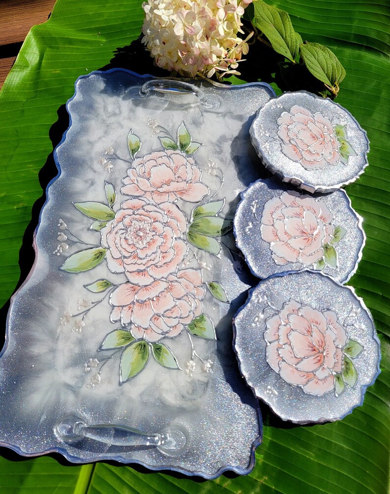 May include: A set of three silver glitter resin coasters with a white background and hand-painted pink peonies. The coasters are round and have a scalloped edge. The tray is also silver glitter resin with a white background and hand-painted pink peonies. The tray has two handles.