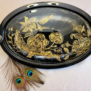 May include: Black oval tray with gold floral design and two gold knobs. The tray is on a white surface with two peacock feathers.