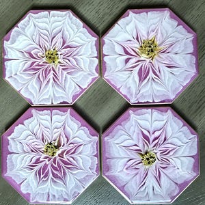 May include: Four octagonal coasters with a floral design. Each coaster features a white and purple flower with a gold center. The petals have a textured, layered appearance, and the background is a solid purple.