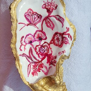 May include: A white oyster shell with a gold rim, painted with pink flowers and a butterfly. The shell is a decorative object.