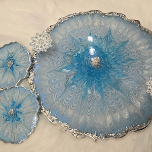 May include: A decorative resin tray and coasters set. The tray and coasters have a blue and white swirl design with silver glitter accents. The edges are trimmed with silver glitter. Two silver snowflake embellishments are attached to the tray.