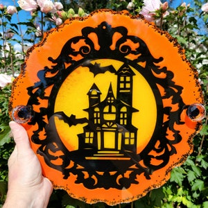 May include: Orange and black Halloween-themed wall decor with a silhouette of a haunted house in front of a full moon. The house has a black silhouette with white windows and a door. The moon is yellow with a black bat flying in front of it.