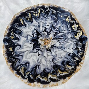 May include: Round resin tray with a swirling design in black, white, and gray. The edges are trimmed with gold leaf and have a textured, organic shape. Two dark blue crystal-like embellishments are on the sides.