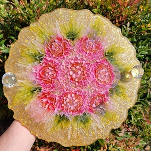 May include: A round, gold resin tray with a pink and green floral design. The tray has a glittery finish and is decorated with two crystal-like embellishments.
