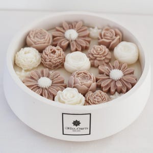 Puede incluir: Un portavelas redondo blanco lleno de velas con forma de flor en tonos blancos y marrones. Las velas están dispuestas para parecer un arreglo floral. El portavelas tiene una etiqueta que dice "Mesa Crafts England."