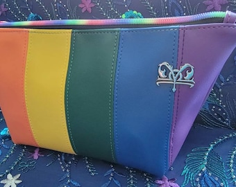 May include: A rainbow-striped cosmetic bag with a zipper closure. The bag features a color gradient of red, orange, yellow, green, blue, and purple. A silver emblem is on the blue panel. The zipper pull is rainbow-colored.