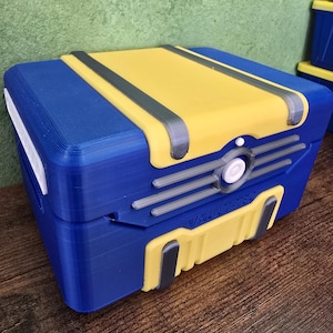 May include: A blue and yellow rectangular box with the text "VAULT-TEC". The box has a yellow top with gray accents and a white circular button. The sides are blue with yellow and gray details.