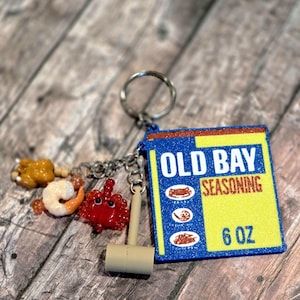 May include: A keychain featuring a rectangular charm designed like an Old Bay Seasoning 6 oz tin with blue, yellow, and red colors. Attached are miniature charms of a crab, a shrimp, a chicken leg, and a small beige mallet on a metal ring.