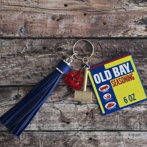 May include: A keychain featuring a blue tassel, a red crab charm, a miniature wooden mallet, and a small rectangular patch with "OLD BAY SEASONING" and "6 OZ" printed on it. The keychain is on a silver ring.