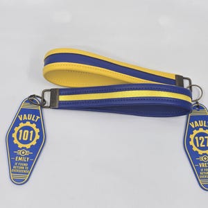 May include: Two wrist lanyards in blue and yellow with attached motel-style key tags. One tag reads VAULT 101 EMILY IF FOUND RETURN TO OVERSEER, the other reads VAULT 127 VREXA IF FOUND RETURN TO OVERSEER. The lanyards feature a blue and yellow color scheme.