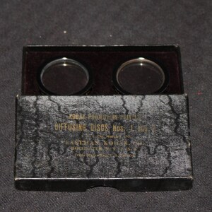 May include: A black box with two round, black diffusing discs inside. The box has gold lettering that reads "Kodak Protection Filter Diffusing Discs No. 1 Fastax Kodak"