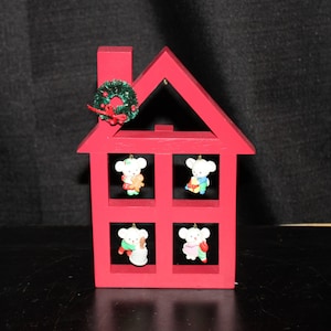 May include: A red wooden house-shaped Christmas decoration. The house has four windows, each with a small mouse ornament. A green wreath with a red bow adorns the roof. A festive holiday decoration.
