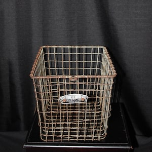 May include: A vintage, rectangular wire basket with a rusted, aged appearance. The basket has a grid-like design and a small label that reads "61 AMERICAN". The basket is sitting on a black surface against a dark background.