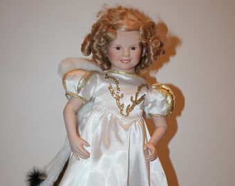 Pode incluir: Uma boneca de porcelana com cabelo loiro encaracolado vestindo um vestido de cetim branco com detalhes dourados. A boneca tem um xale de pele branca e está em pose com um sorriso doce.