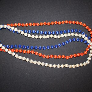 May include: Three strands of beaded necklaces in red, white, and blue, arranged on a black surface. The beads are small and round, with a satin-like sheen. The necklaces are intertwined, creating a patriotic color scheme.