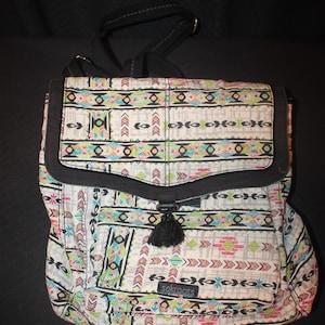 May include: A patterned backpack with a black flap and strap. The bag features a white background with a colorful geometric design in pink, teal, and green. A black tassel hangs from the flap. The brand name "sakroots" is visible on the front.