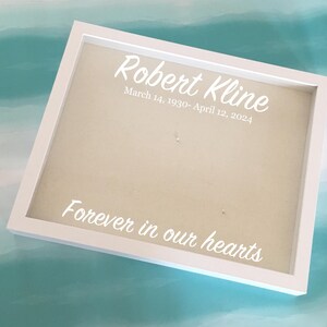 Puede incluir: Caja de sombra enmarcada en blanco con el texto "Robert Kline, March 14, 1930-April 12, 2024" y "Forever in our hearts" en escritura blanca. El fondo es de color beige claro.
