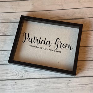 Puede incluir: Caja de sombra enmarcada en negro con el nombre "Patricia Green" y las fechas 13 de noviembre de 1945 - 3 de junio de 2025 en fuente cursiva. El fondo es de tela beige claro.