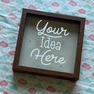 Puede incluir: Una caja de sombra cuadrada de madera marrón oscuro con las palabras "Your Idea Here" en escritura blanca. El fondo es de tela gris claro. La caja está sobre una tela estampada con nubes y las palabras "Baby" y "Dream".