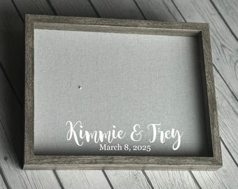 May include: A gray wooden shadow box with a light gray fabric interior. The names "Kimmie & Trey" and the date "March 8, 2025" are written in white script across the bottom of the fabric. The frame has a rustic, weathered appearance.