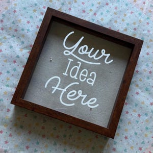 Puede incluir: Un marco de madera marrón oscuro cuadrado con las palabras "Your Idea Here" en escritura blanca. El fondo es una tela beige claro. El marco se muestra sobre una superficie estampada con estrellas.