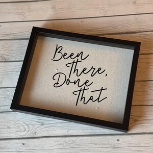 Könnte beinhalten: Ein gerahmtes Schild mit dem Schriftzug "Been There, Done That" in eleganter schwarzer Schrift. Das Schild hat einen neutralfarbenen Hintergrund und ist in einem schwarzen rechteckigen Rahmen gefasst, geeignet für die Inneneinrichtung.