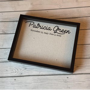 Puede incluir: Una caja de sombra negra con el nombre "Patricia Green" y las fechas "November 13, 1945- June 3, 2025" impresas en escritura negra. La caja tiene un respaldo de tela de color claro y se muestra sobre una superficie de madera clara.