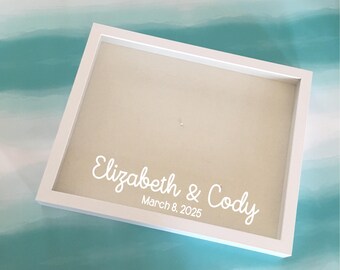 May include: White shadow box with a beige interior. The text "Elizabeth & Cody" and "March 8, 2025" are written in white script. The box is angled, set against a light blue and white watercolor background.