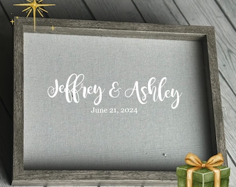 May include: A framed gray canvas with the names "Jeffrey & Ashley" and the date "June 21, 2024" in white script. A small green gift box with a gold bow sits in the corner. The frame is a weathered gray color.