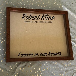 Puede incluir: Un memorial enmarcado con el nombre "Robert Kline" y las fechas "14 de marzo de 1930 - 12 de abril de 2024" en escritura negra. La frase "Forever in our hearts" también se muestra. El marco es de madera marrón oscuro.