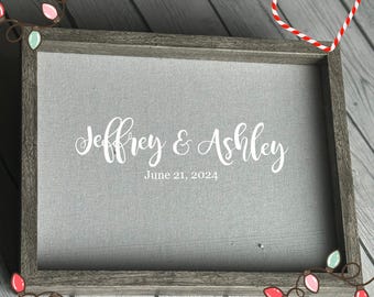 May include: A framed gray canvas with the names "Jeffrey & Ashley" in white script, and the date "June 21, 2024" below. The frame is a weathered gray wood. Decorative string lights and a candy cane heart are in the corners.